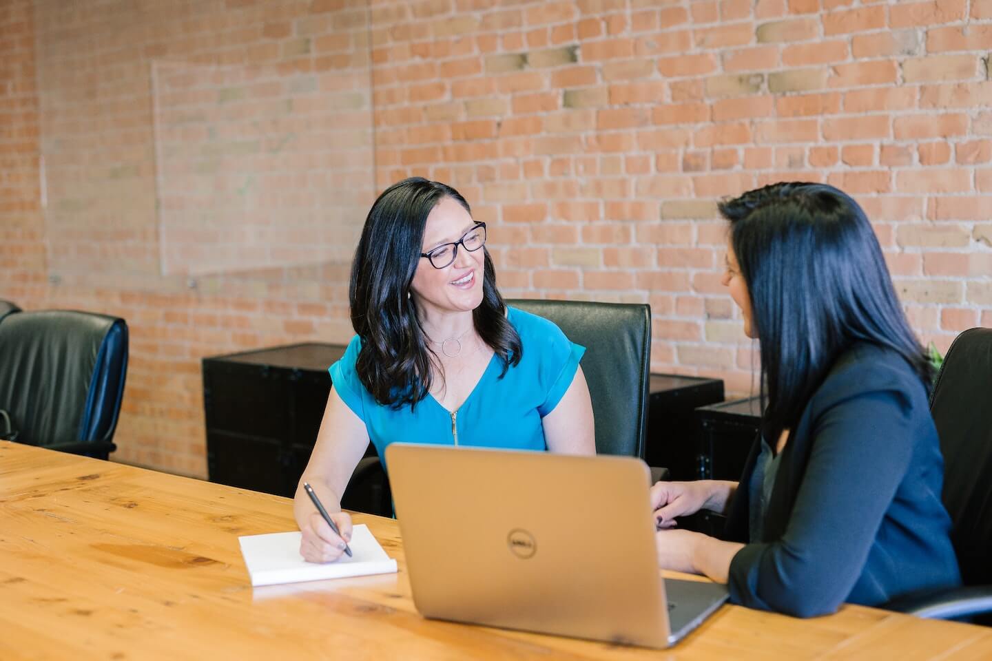 Two women in professional-casual attire sit at a wooden conference table in an office with a brick wall, discussing work with a laptop and notebook.