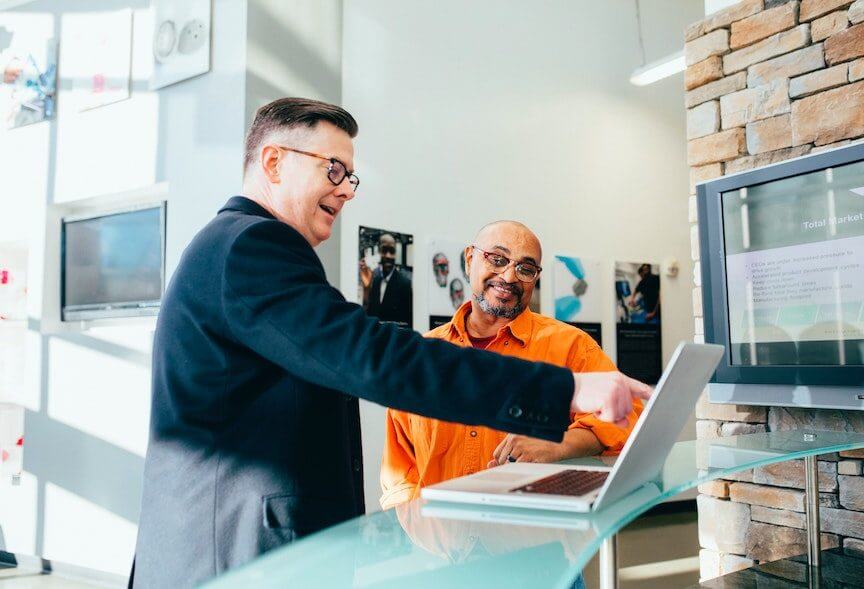 Two men in a modern office discuss content on a laptop screen, with one man pointing at the display while the other looks on and smiles.