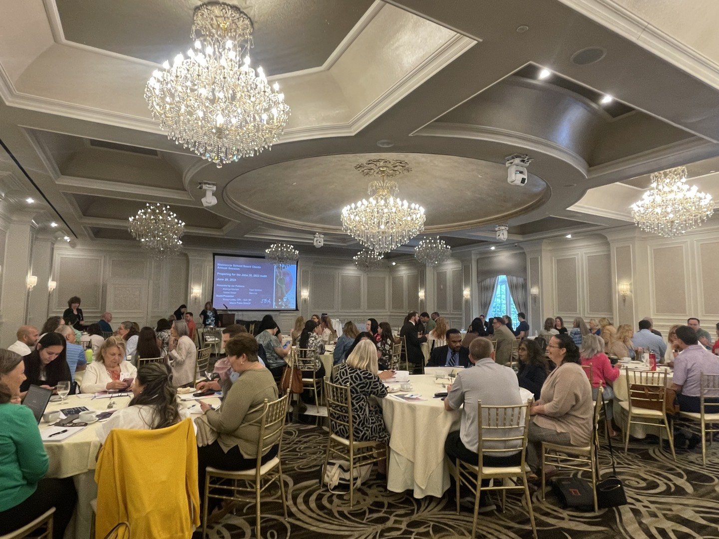 Large ballroom with attendees seated at round tables for a professional event, illuminated by ornate crystal chandeliers.