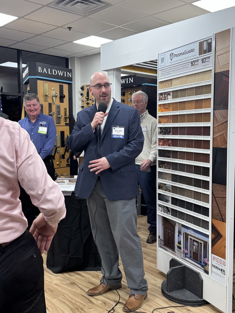 A man in a navy blazer and grey slacks stands in a showroom holding a microphone and addressing a crowd. He wears a "SPEAKER" badge and stands next to a large display of door finish samples. Several other men in professional-casual attire are visible in the background among displays of door hardware.