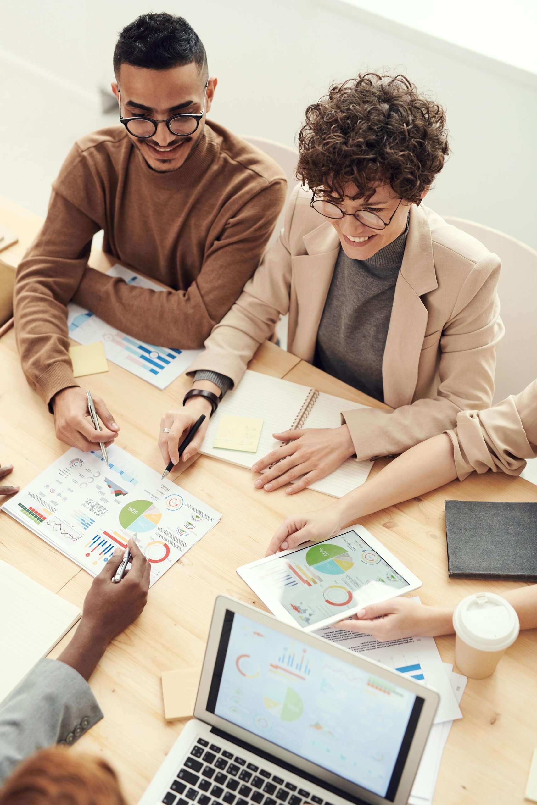 An overhead view of colleagues smiling while reviewing colorful data charts and a tablet during a productive meeting at a wooden table.