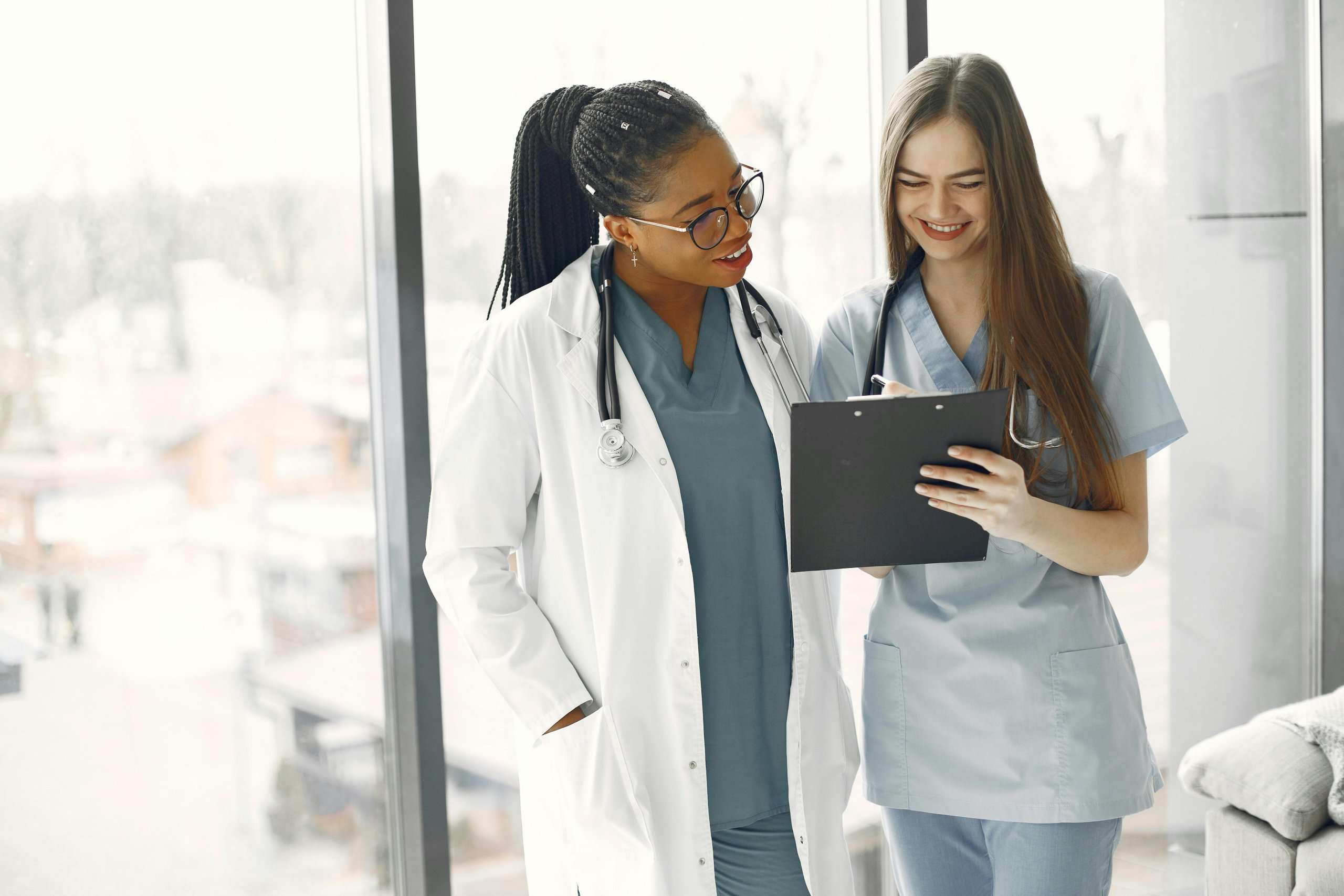 Two healthcare professionals, one in a white lab coat and the other in blue scrubs, smile while reviewing notes on a clipboard in a bright hospital setting.