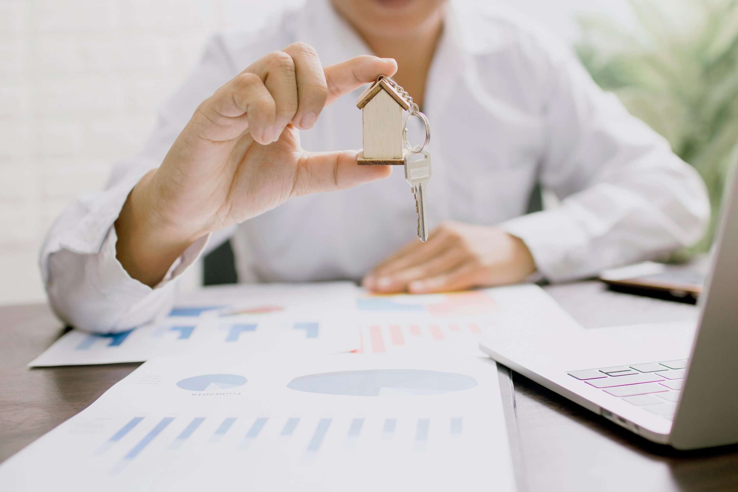 A person in a white shirt holds up a house-shaped keychain with a key over a desk covered in financial charts and graphs.