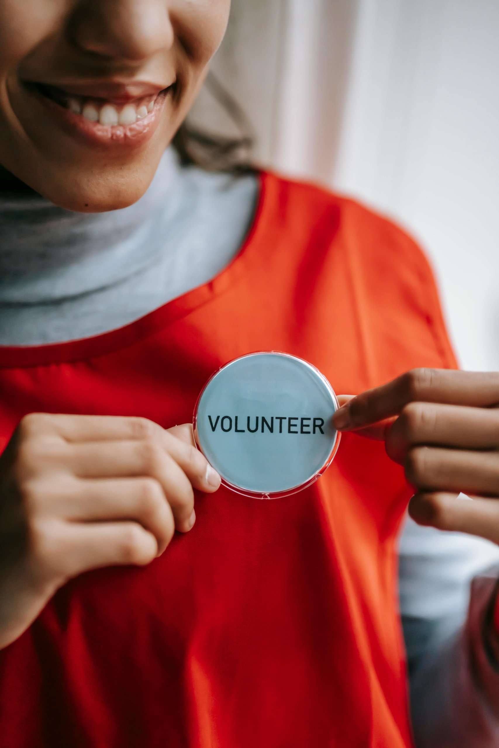 A smiling person wearing an orange vest holds a round blue badge that says "VOLUNTEER" in the center of their chest.