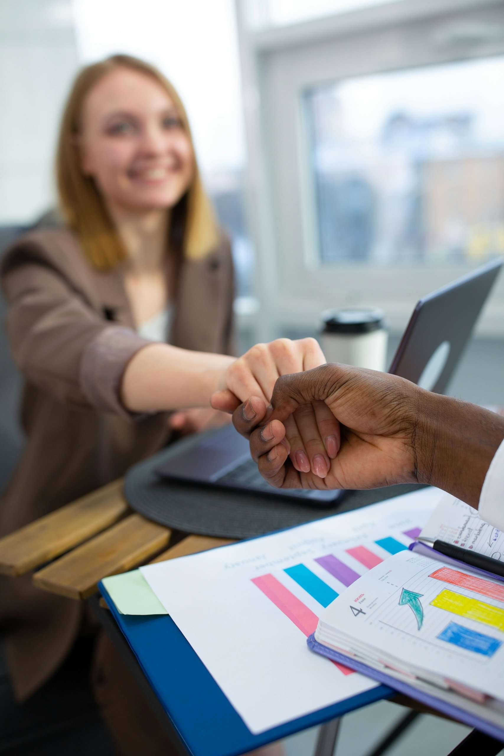 A smiling professional woman shakes hands with a colleague across a desk featuring a laptop, charts, and a colorful data report.