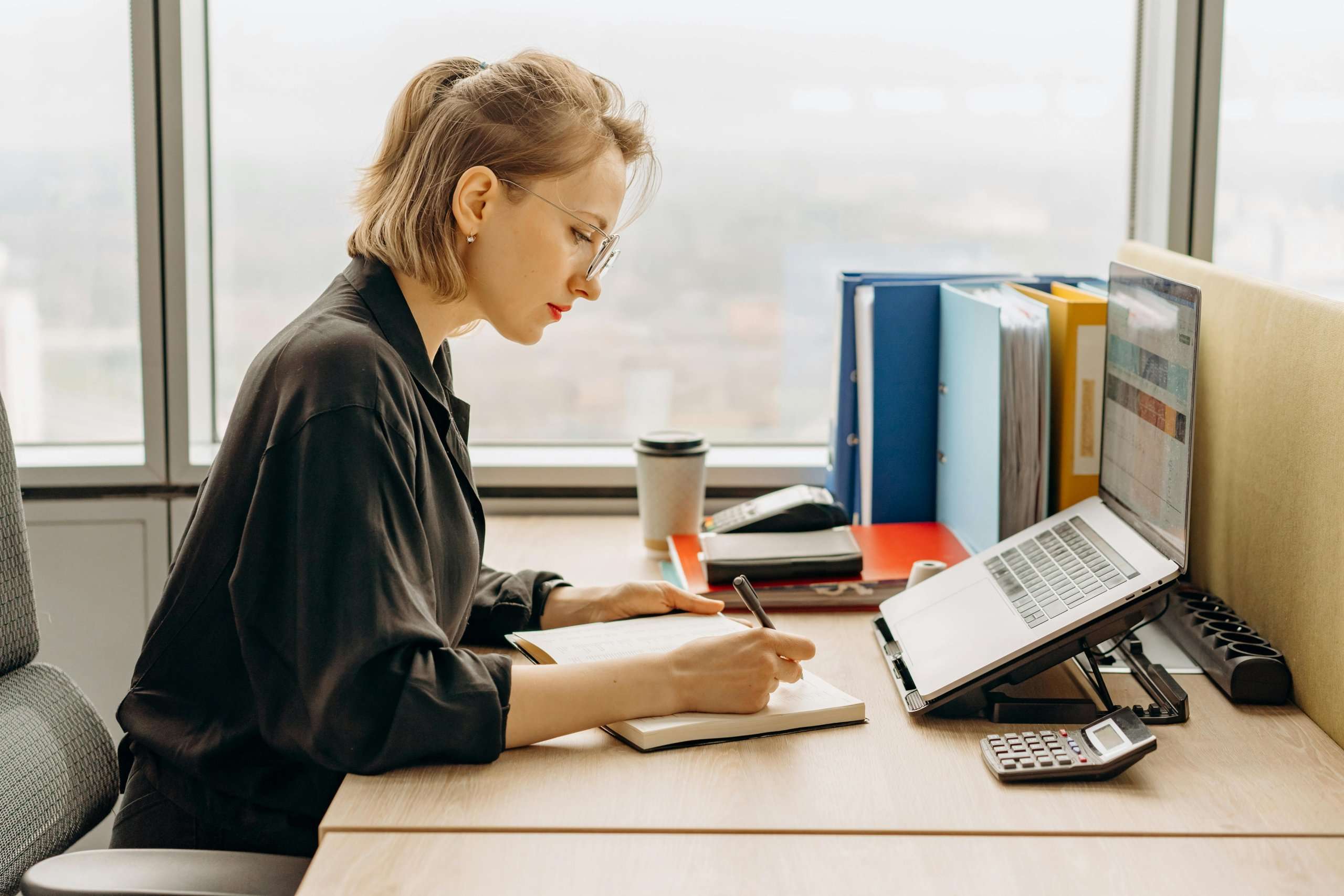 A woman in a black shirt sits at an office desk by a window, writing in a notebook. Her workspace includes a laptop on a stand, a calculator, and colorful binders.