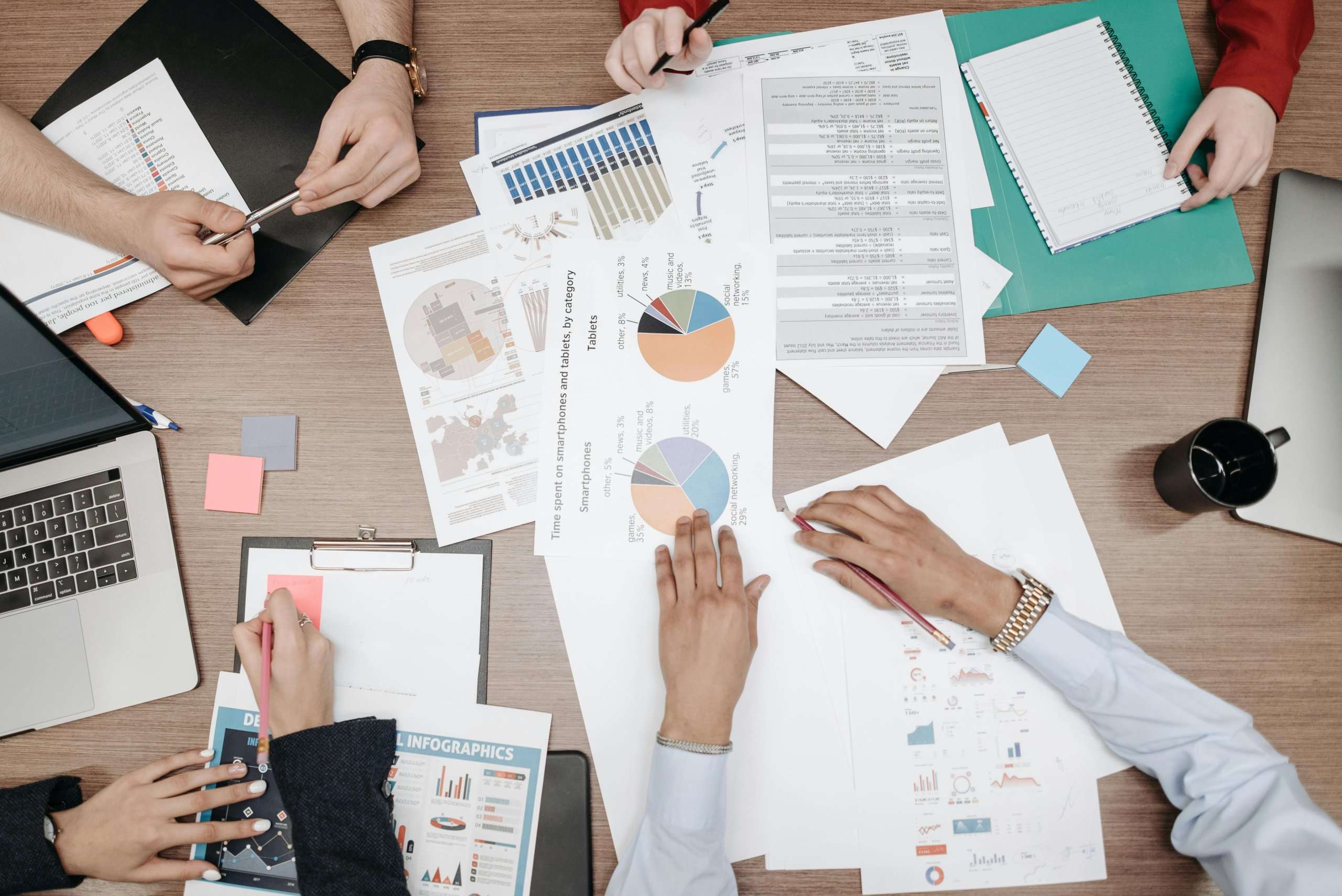 An overhead view of a team collaborating around a wooden table, featuring laptops, notebooks, and papers with colorful pie charts and infographics.