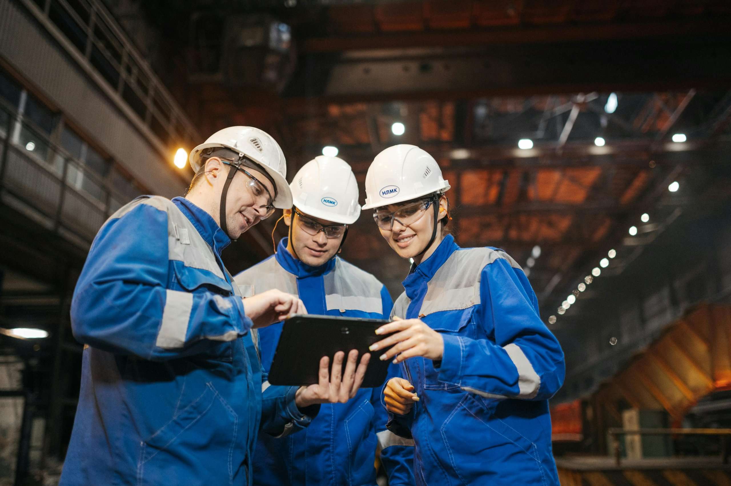 Three industrial workers in blue uniforms and white hard hats collaborate in a large warehouse, looking at a digital tablet together.