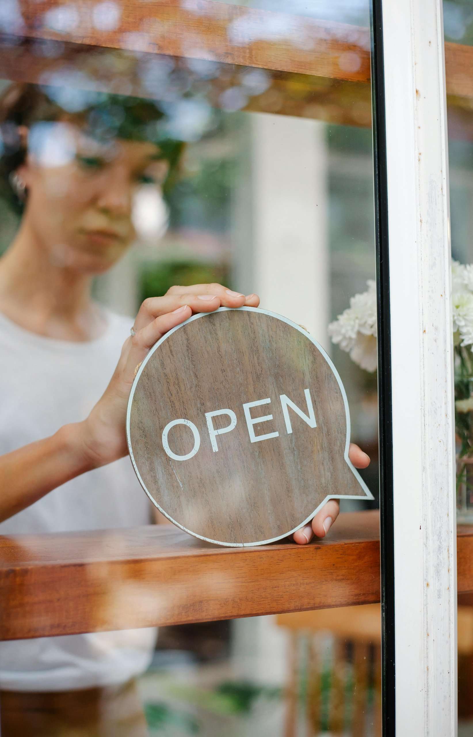 A person behind a glass door hangs a circular wooden "OPEN" sign shaped like a speech bubble.