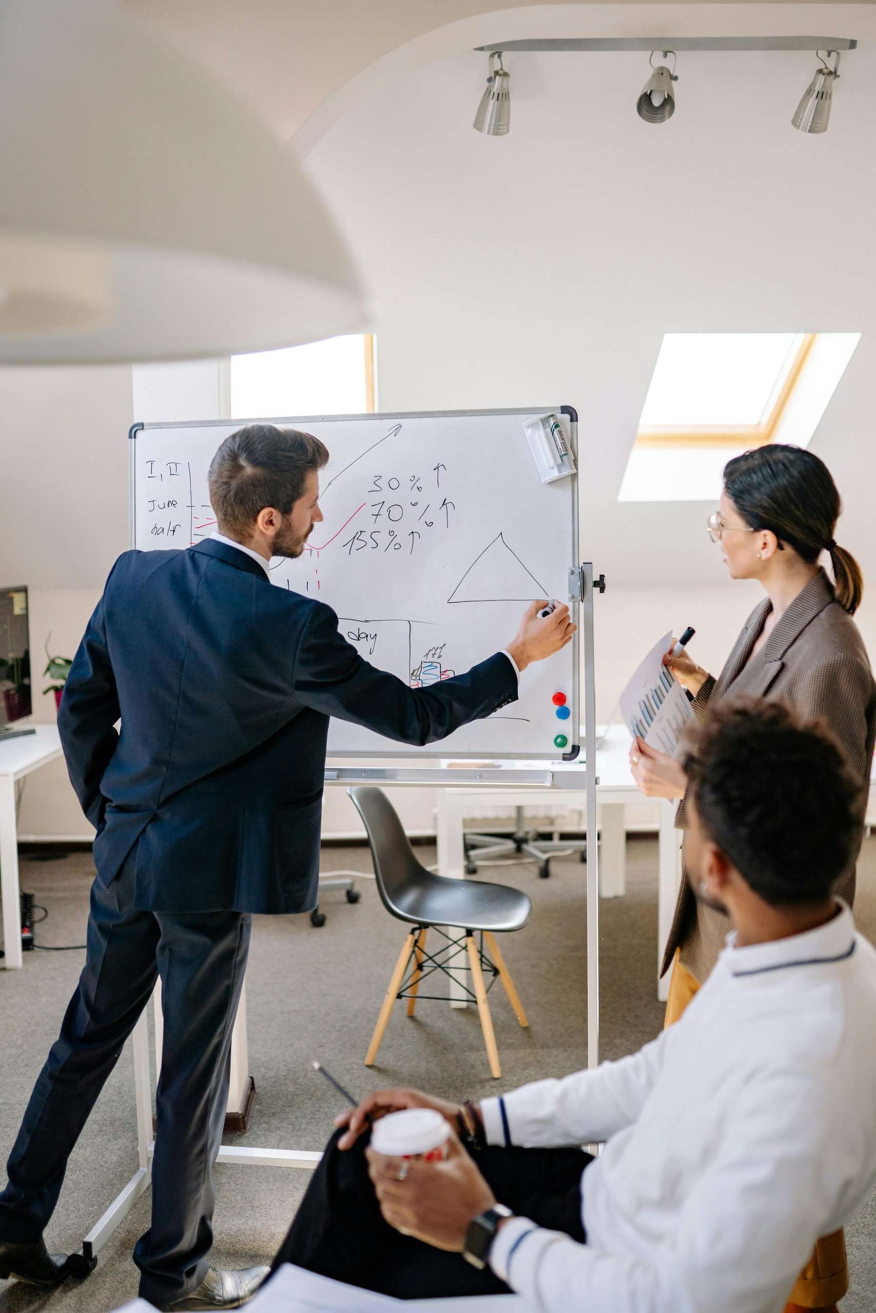 A group of three professionals in business attire collaborate during a presentation in a bright, modern office. A man in a dark suit draws a triangle and data trends on a whiteboard, while a woman beside him holds a report and a seated man observes from the foreground.