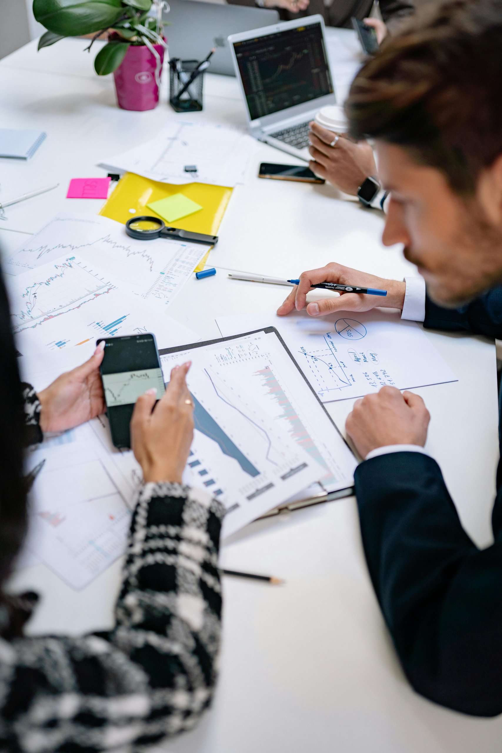 Top-down view of people working at a white desk covered in data charts, sticky notes, and a laptop. One person holds a smartphone showing a graph, while another points at a printed report with a pen.