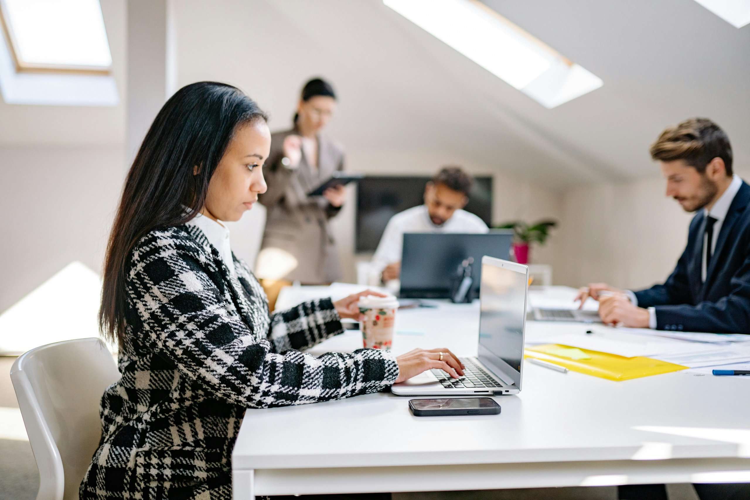 A woman in a plaid blazer works on her laptop at a desk in a collaborative attic office.