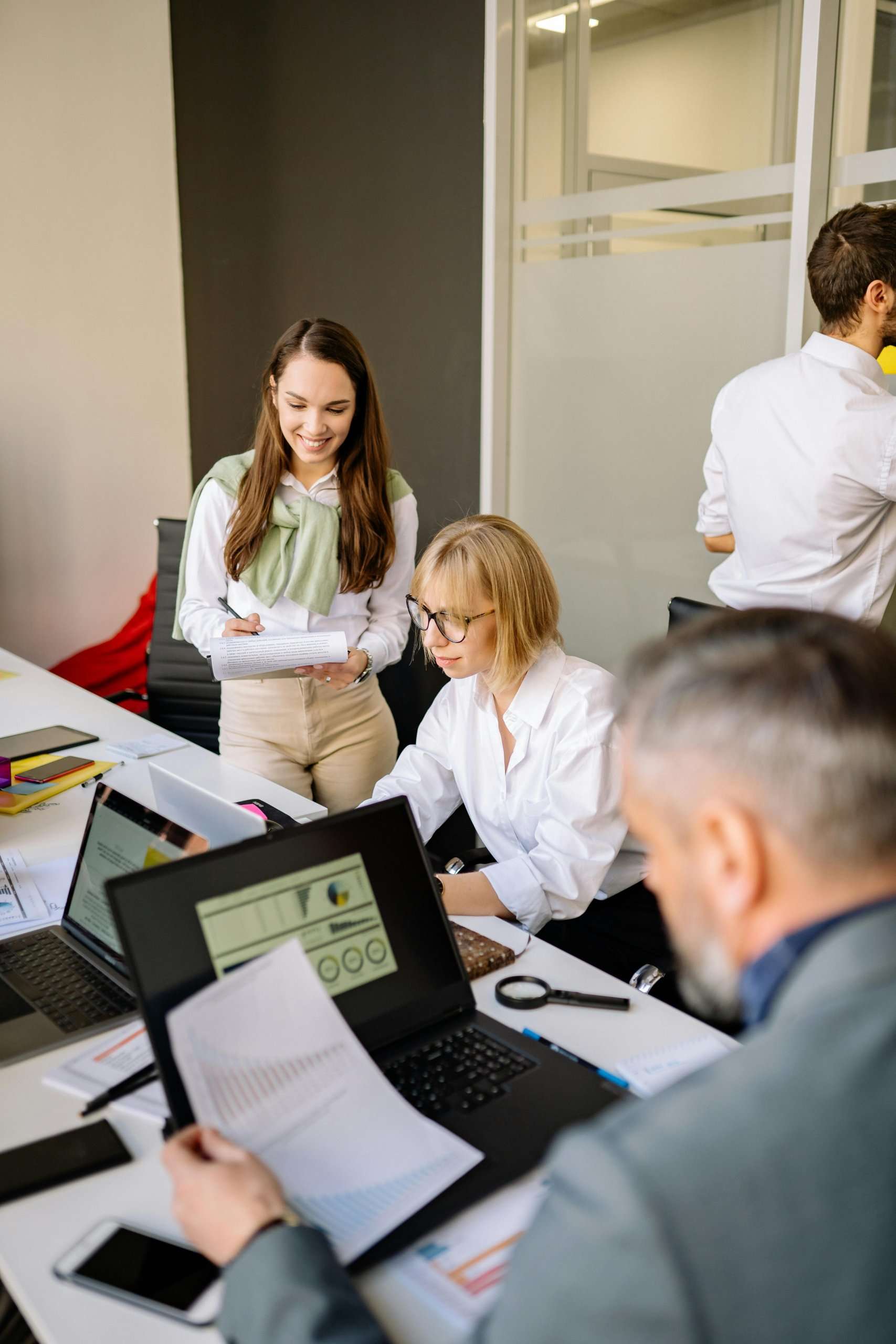 Professionals collaborating at a long desk with laptops and reports in a contemporary office setting.