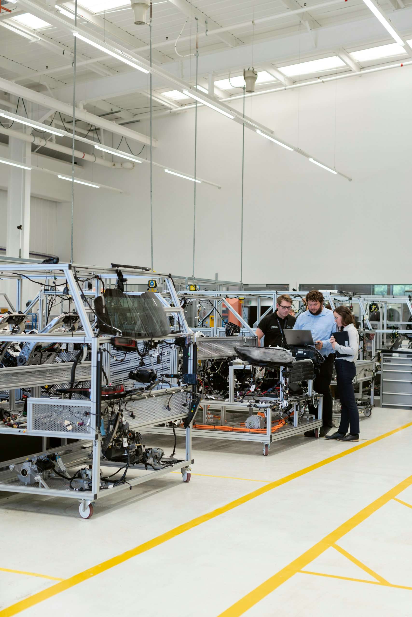Three engineers stand in a bright, modern manufacturing facility, reviewing technical data on a laptop while working on an assembly line with complex mechanical frames.