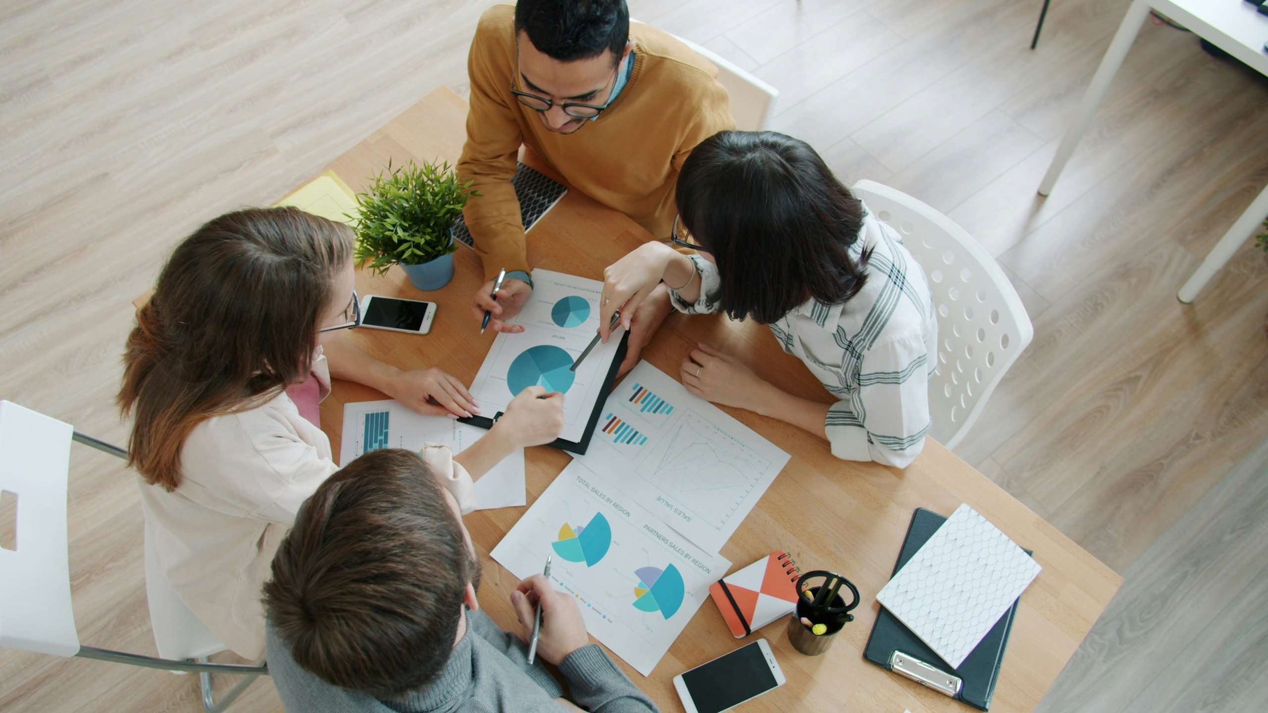 An overhead view of four colleagues gathered around a wooden table, discussing printed pie charts and data reports while taking notes.