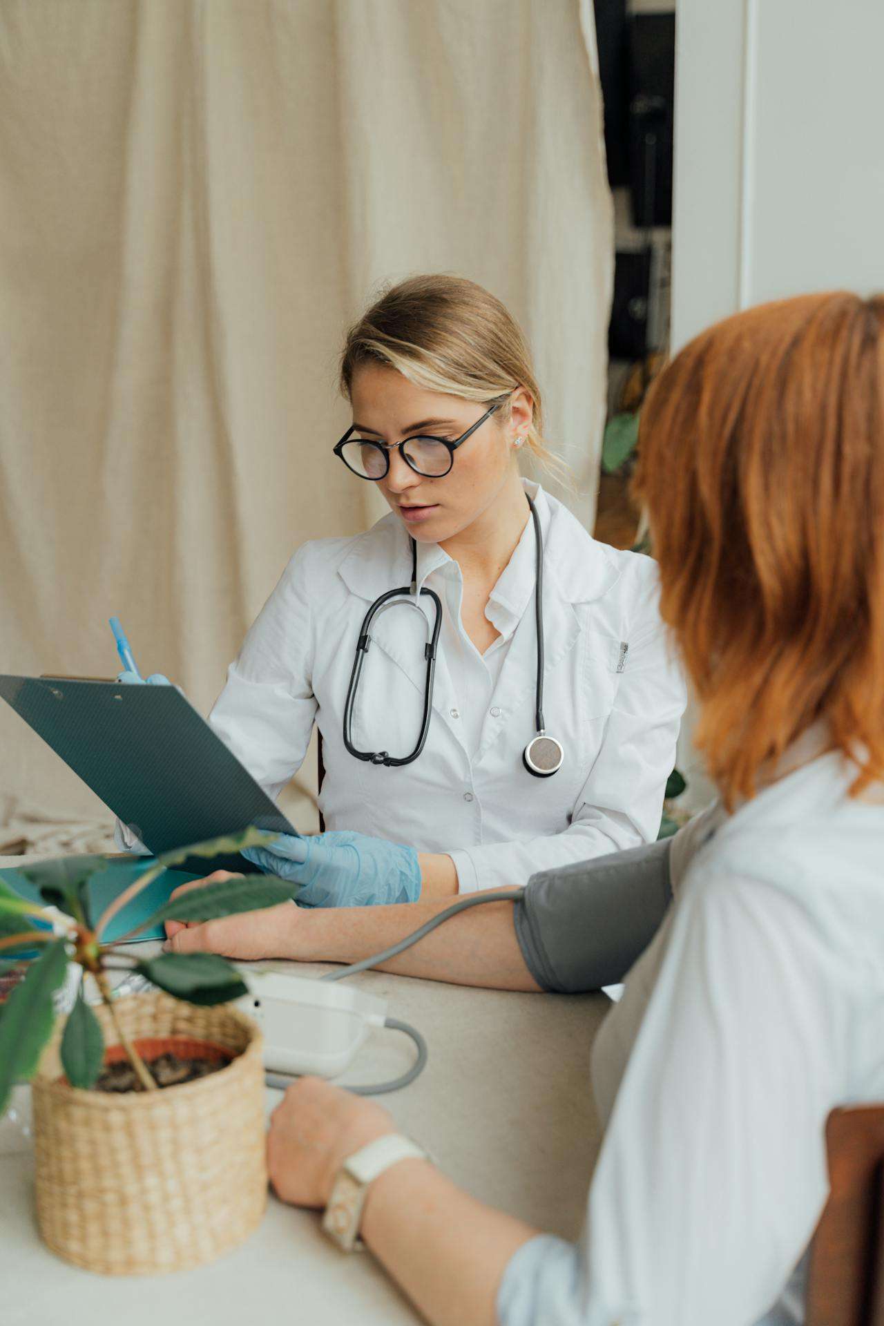 A doctor in a white coat and stethoscope reviews a clipboard while taking a patient's blood pressure during a medical consultation.