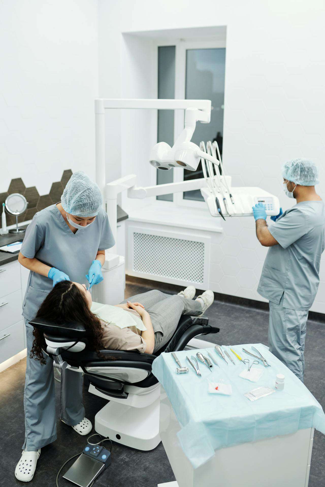 Two dental professionals in scrubs and gloves treating a patient reclined in a dental chair, with instruments laid out on a tray in a modern clinic.