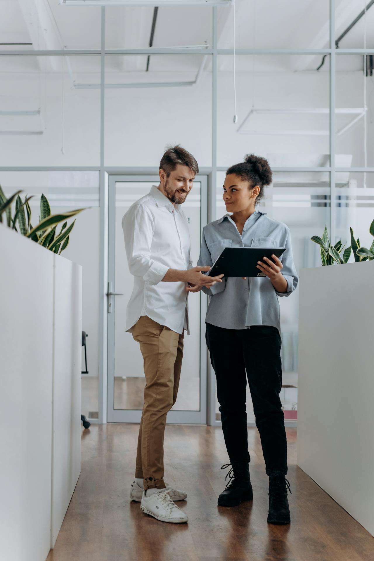 Two coworkers standing in a modern office hallway reviewing documents on a tablet together.