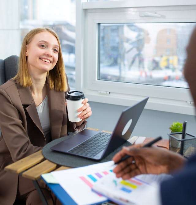 A smiling woman holding a coffee cup listens during a business meeting, with laptops and colorful charts on the table.