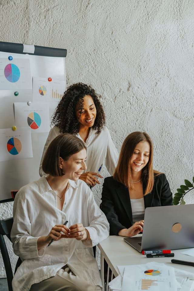 Three women collaborating in an office meeting around a laptop with charts and reports visible.