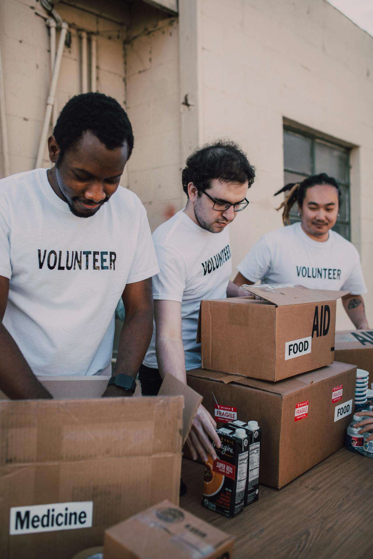 Three volunteers in white t-shirts packing food aid and medicine boxes at a community relief drive.