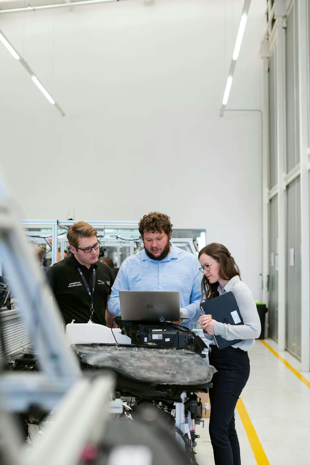 Three engineers, including a Jaguar apprentice, gathered around a laptop on an automotive assembly line in a manufacturing facility.