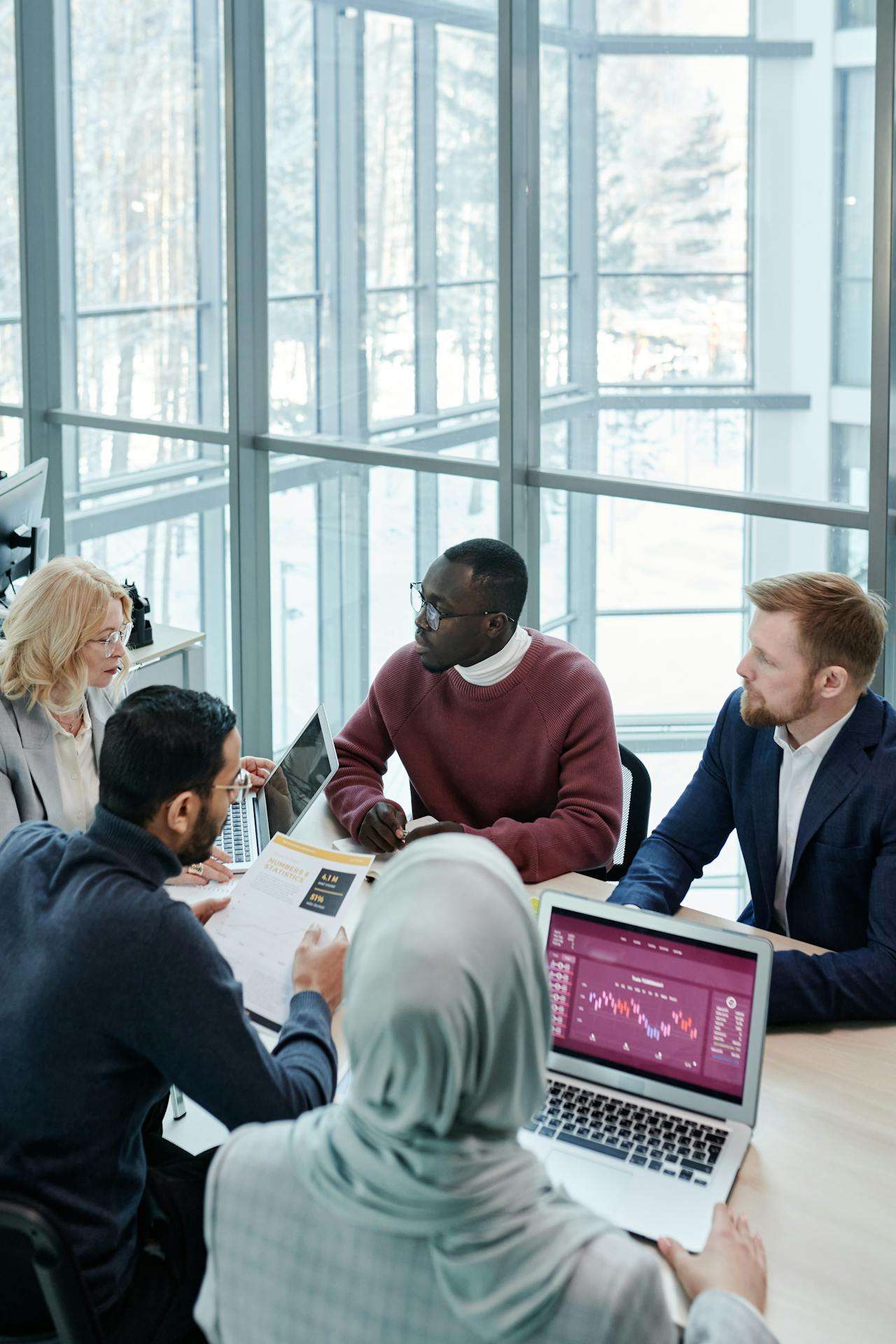 A diverse team of five professionals in discussion around a conference table with laptops and printed reports, in a bright glass-walled meeting room.