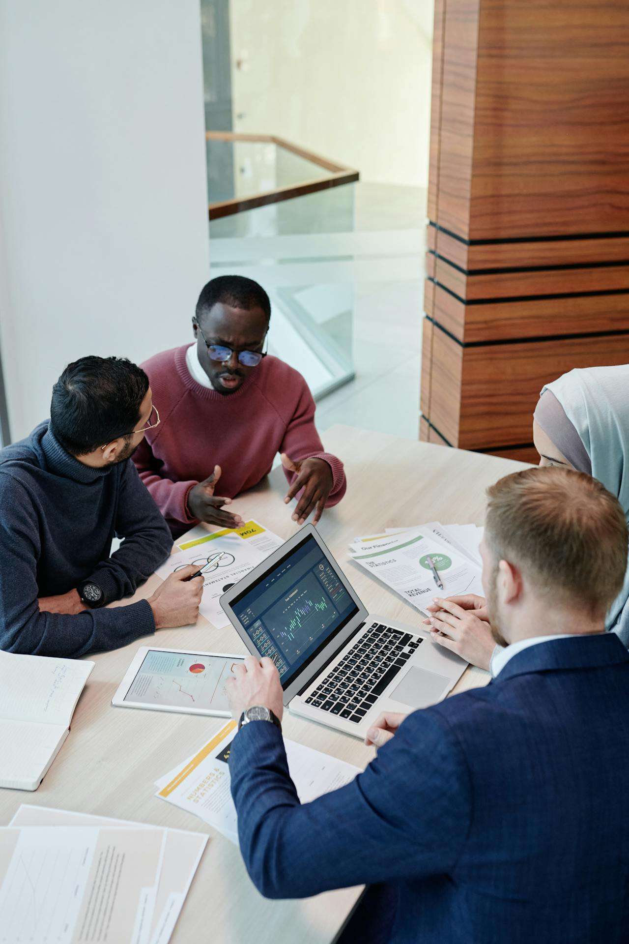 A diverse group of four colleagues collaborating around a table with a laptop showing financial charts, a tablet, and printed reports during a business meeting.