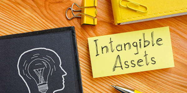 yellow sticky note on table with 'intangible assets' written in pen, next to pen, paper clip, notebook, and black folder with white graphic of a side profile and lightning bulb drawn in ink