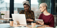 Two women collaborate in a modern office, focused on a laptop and a tablet at a wooden desk with coffee cups. One wears a patterned shirt and headwrap, while the other has short blonde hair and wears a maroon top.